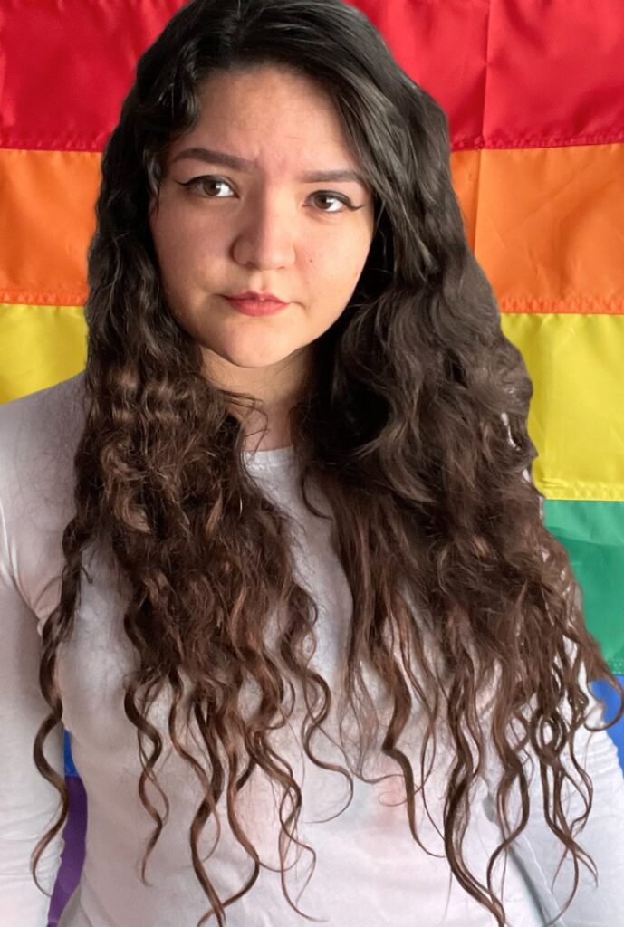 Young woman with long brown curly hair standing in front of a rainbow flag